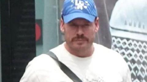 Ryan Wedding in a white T-shirt and blue baseball cap, with a black bag strap across his shoulder. 