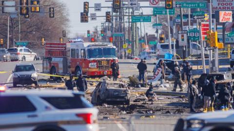 the scene of the crash, showing burnt out debris, cars and a fire truck in the background