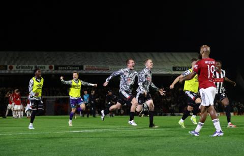 Manchester United's Bryan Mbeumo walks away after missing the final penalty. Grimsby players celebrate wildly after winning and are running to hug one another as other Manchester United players hold their heads in the background. The sky is dark as the game finished late after the penalty shootout