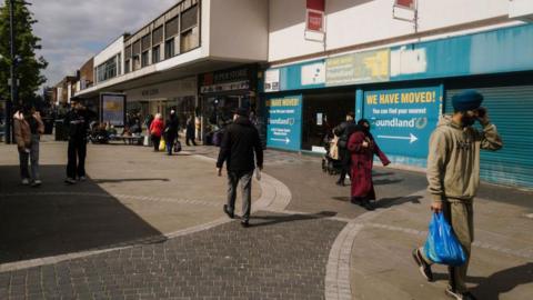 A picture depicting West Bromwich High Street during the day, showing a closed Poundland store and shoppers in the background. A man in the foreground is dressed in a beige tracksuit and turban and carrying a blue plastic bag.