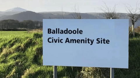 A white sign which reads "Balladoole Civic Amenity Site" in black text, with a grassy green field and hills in the background. 