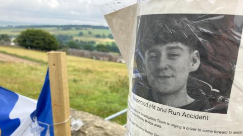 A memorial-style notice is taped to a wooden pole in a rural roadside setting. The notice includes a photo of a young man and text about a suspected hit‑and‑run near Pateley Bridge. Behind the pole are open fields, trees, and distant hills under a bright sky. A blue‑and‑white flag and a string of small lights hang nearby.