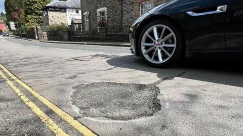 A pothole on Dolvin Road in Tavistock with a passing navy blue car.