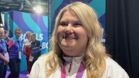 A lady with blonde shoulder-length hair is wearing a white jacket with a purple and blue medal ribbon around her neck