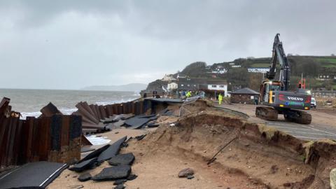 Engineers work on a road which has partially collapsed along Devon's coast. A digger is parked next to the area which has collapsed.