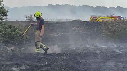 A single firefighter walking around a blackened field with wisps of smoke coming off it and a fire engine in the background