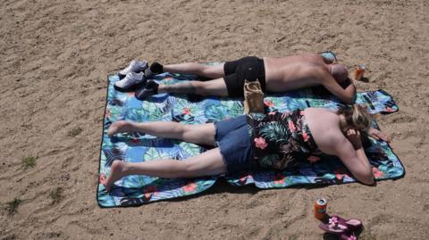 A man and woman lie face down on a mat, on a sandy beach, with their backs exposed to the sun. Two cans of orange fizzy drink are sitting on the sand.