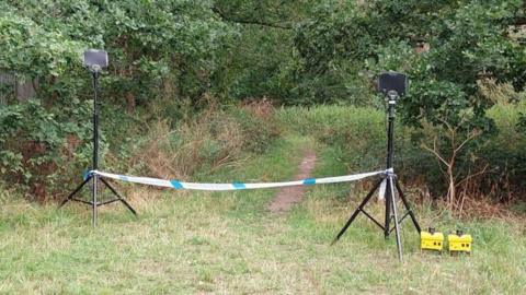 A police cordon in place in a field, with trees beyond it.