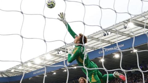 Mads Hermansen dives upwards to try to reach the ball after Jhon Duran's header. The ball is looping over his outstretched right arm with the picture being taken from behind the bet, with the netting visible in the foreground. 