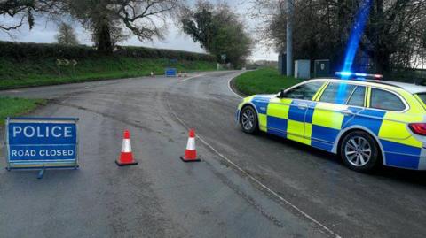 A bend on a country lane, curving round to the right. It is edged by grass verges and a hedge and trees. A police car - blue and neon - is parked on the right hand carriageway. There are dirt tyre-marks across the road. There are two blue signs saying "Police ROAD CLOSED" and four traffic cones blocking the road.