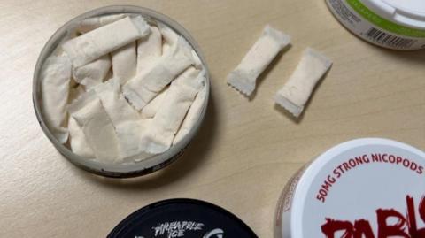 An open tin of nicotine pouches on a wooden table. There are two pouches out of the tin and two unopened tins on the side. One says 50MG Strong Nicopods on the lid.