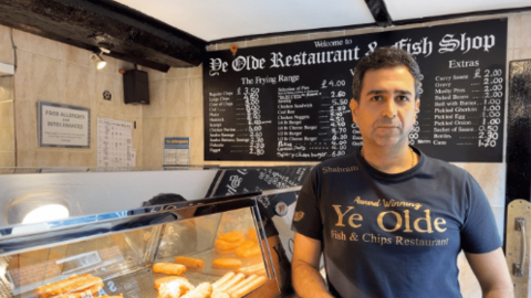 Shahram Navard looks into the camera with a solemn expression. He is wearing a navy t-shirt with Ye Olde Fish and Chips Restaurant written on it. He is leaning against a glass counter where food is visible. Behind him is a blackboard with prices written in chalk.