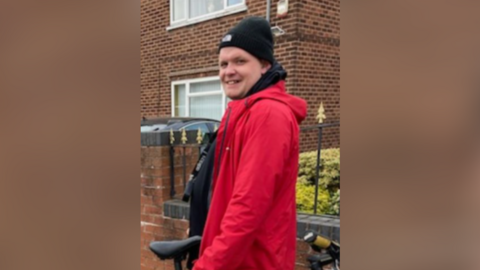 A man wearing a red coat and black woolen hat.  He is leaning on a bike