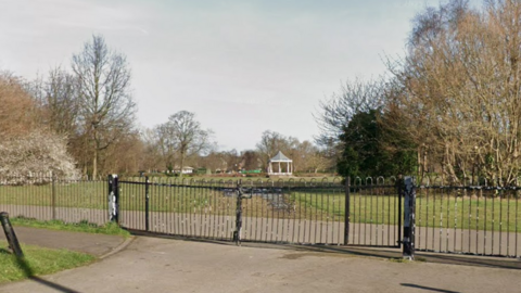 A view of Albert Park over the railings. A footpath is close to the gates and there is a bandstand in the background.