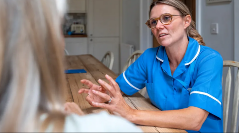 A nurse talking to a patient at home. We see the blonde hair of the patient and the front of the nurse who wears a blue uniform, has glasses and long hair swept back behind her ears. They sit at a wooden table