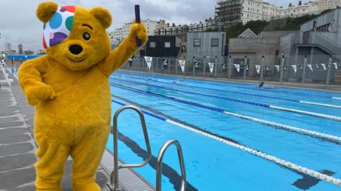 Pudsey, the Children in Need mascot, holding a relay baton standing next to a swimming pool