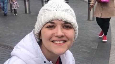 Jo Broadhurst seen in the city centre with shoppers around her. She is in a pedestrian area and there are bollards and shops in the background. She is wearing a white puffer jacket and a white woolly hat and is smiling at the camera.