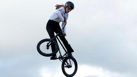 A woman wearing a black helmet, white top and black trousers, is mid-air while she performs a trick on a BMX.