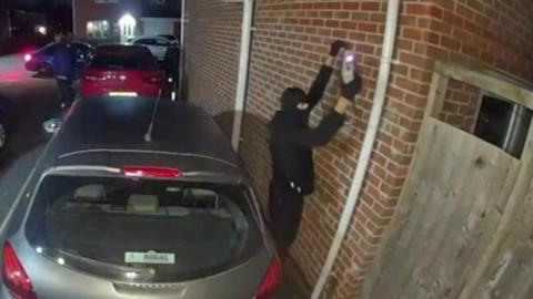 A CCTV image showing a person dressed in black clothing and wearing a balaclava holding a device against a brick wall. The individual is standing in a residential driveway next to a silver car parked close to the wall, with other vehicles visible in the background under street lighting. A wooden fence is partially visible on the right side.