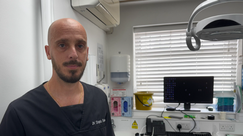 Dr Dario Sales, who has a shaved head with a brown beard and moustache, is photographed wearing a black tunic in his dental surgery.