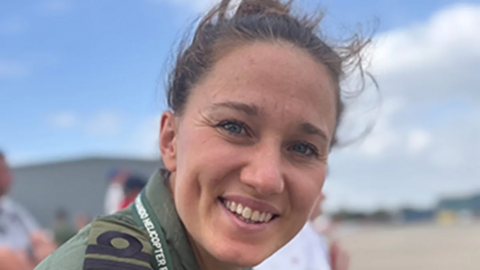 A picture of Lily-Mae Fisher, who is smiling to the camera with her hair tied back and windswept. She is wearing a green shirt for her uniform and a lanyard that says "helicopter" on it.