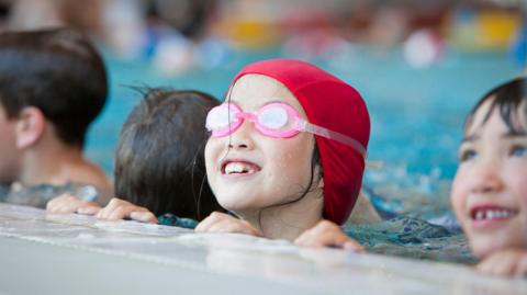 A young girl wearing a red cap and pink goggles in the water at the edge of a swimming pool during a swimming lesson. There are three children next to her.