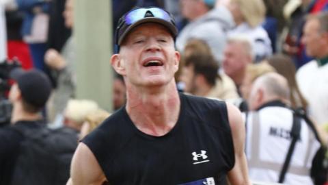 A man is running in a black and orange tank top, with a black hat and blue sunglasses on his head. He has an exhausted expression and in the background there is a large crowd of people behind a barricade.