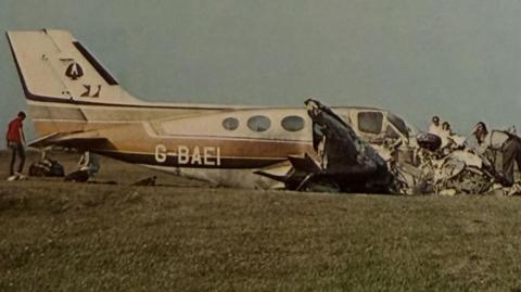 An old photograph shows the wreckage of a small aircraft in a grassy field while people dig through its remains and others watch on.
