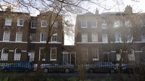 A row of terraced houses, all of which are three storeys high apart from number 25 and 26 which stand at one storey with their front doors next to each other.