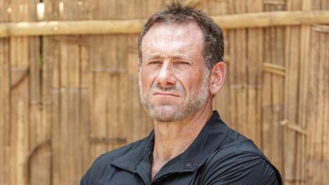 A publicity photo of Jason 'Foxy' Fox wearing a black shirt. He has a grey beard and short brown hair. He is pulling an intimidating face.