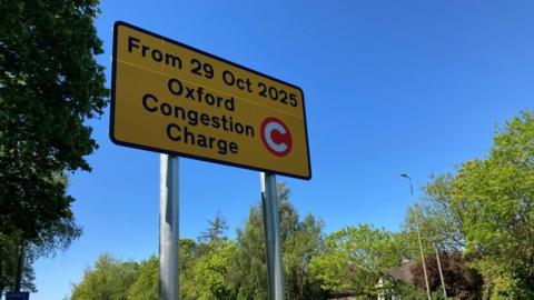 A yellow sign, high in the sky. It says "From 29 Oct 2025, Oxford Congestion Charge" alongside a white C in a red circle.