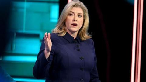 Penny Mordaunt in a navy blue suit, looking to her left with arm and hand extended. She has shoulder-length blonde hair