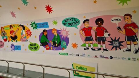 A portion of the mural. It covers part of the enclosed bridge between the interchange and station. Cartoon images of children from different backgrounds are depicted playing with speech bubbles of northern slang - such as "nah then" and "mucka". They are accompanied by information about local support services and QR codes.