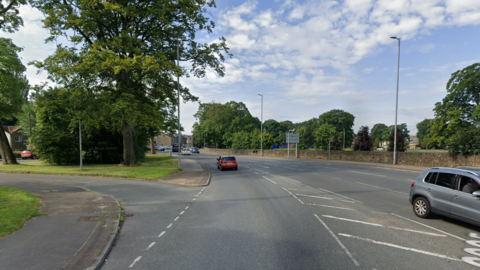 A stretch of road sweeps to the left. Two cars can be seen. On one side of the road is a stone wall.