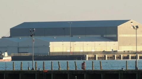 An image of grey, industrial buildings based at the Arnish fabrication yard near Stornoway. The picture is taking from across a body of water, and sunlight shines against the side of the buildings.