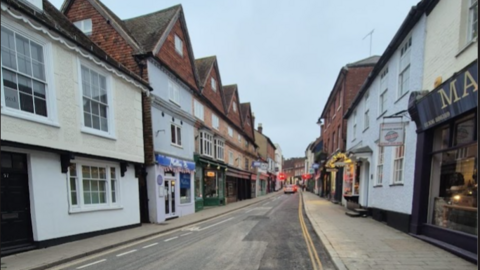 A view down West Street with cars seen in the distance and shops either side. There is a line down the middle of the road where a new surface has been laid on one side.