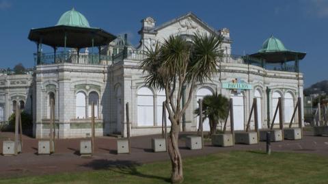 The exterior of Torquay Pavilion which is a white building in a Victorian style. There are two trees in front of it.