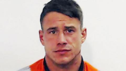 A police mugshot of Cameron McGregor, a man in his early 30s, with short brown hair and wearing an orange and black top