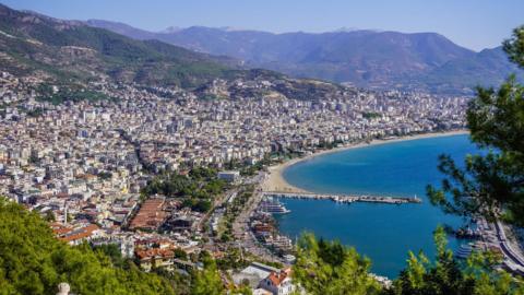 A general view of Alanya in Turkey, which has a beach, large blue sea and lots of buildings. A pier stretches from the beach out to sea.