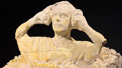 A sculpture of a man putting on ear defenders is made out of plaster. 