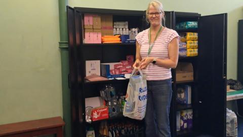 A woman holds a plastic carrier bag containing toiletries. Behind her are shelves stacked with multi-packs of sanitary towels, deodorant, nappies and other hygiene items.