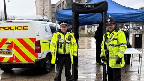 Two male police officers in high-visibility uniforms stand either side of a metal detecting archway set up in a public square. Behind them is a large blue marquee and a police van is parked next to the arch