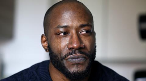 A man with a moustache, beard, silver nose ring and black earring looks away from the camera with tears in his eyes. One of his cheeks is tear-stained and another has a tear rolling down it. He wears a navy blue jumper.