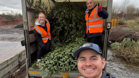 Three people at the back of a van filled with Christmas trees look to the camera, smiling. They are wearing dark clothes with orange high-vis jackets on. There are more Christmas trees on the ground