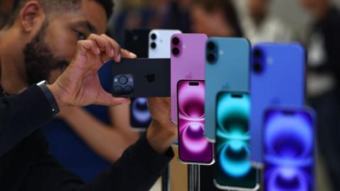 Man dressed in black takes a photo on his smartphone of three Apple iPhone 16s in different colours including plum, teal and midnight blue.