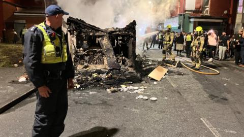 A police officer stands in front of a burnt-out bus