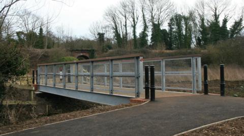 Metal and wooden bridge over a river, hills and trees in the background. A tarmac path leads up to the bridge and there are bollards on both sides