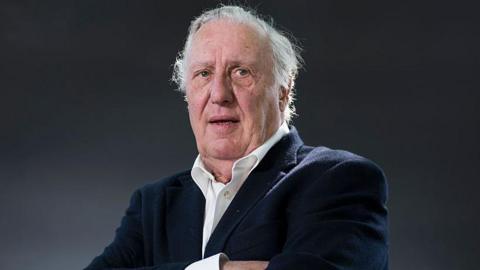 Frederick Forsyth is seen in later life.  He is looking slightly off camera in a head and shoulders portrait. His arms are folded and he is wearing a white shirt and blue jacket.