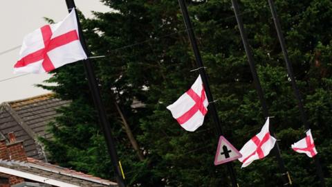 St George's flags fly from a row of black lampposts and are rippling in the breeze. Rooves of nearby buildings can be seen to the left and there are trees in the background