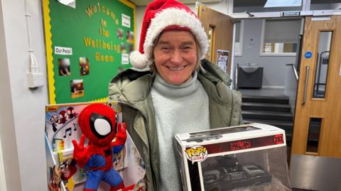 A woman wearing a red Santa hat smiles as she stands in a school corridor holding a boxed singing Spiderman doll and a toy Batman car